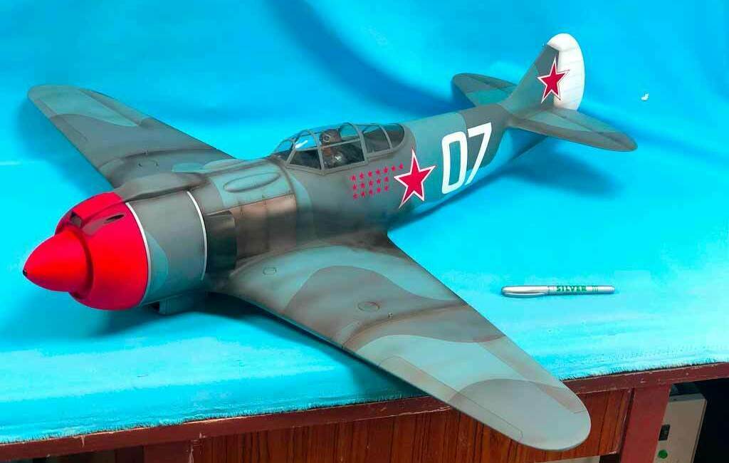 Масштабная модель советственного истребителя с красным носом и звёздами на голубом фоне детализированной масштабной модели советского военного самолёта‑истребителя с камуфляжной раскраской, красным носом и бортовым номером 07, стоящей на столе на фоне голубого экранадетализированной масштабной модели советского военного самолёта‑истребителя с камуфляжной раскраской, красным носом и бортовым номером 07, стоящей на столе на фоне голубого экрана