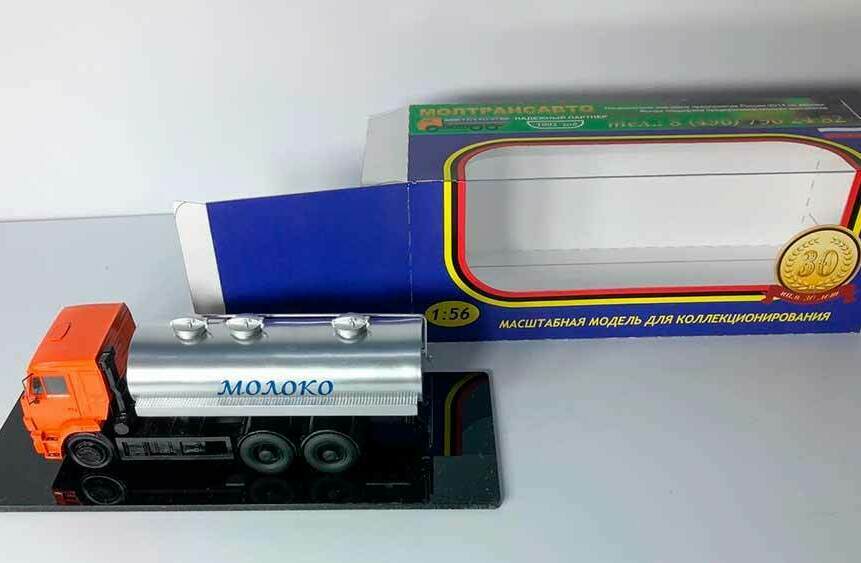 Масштабная модель молоковоза 1:43 в фирменной коробке Коллекционная модель молоковоза в масштабе 1:43 — оранжевый грузовик с серебристой цистерной «Молоко» на подставке рядом с упаковкой