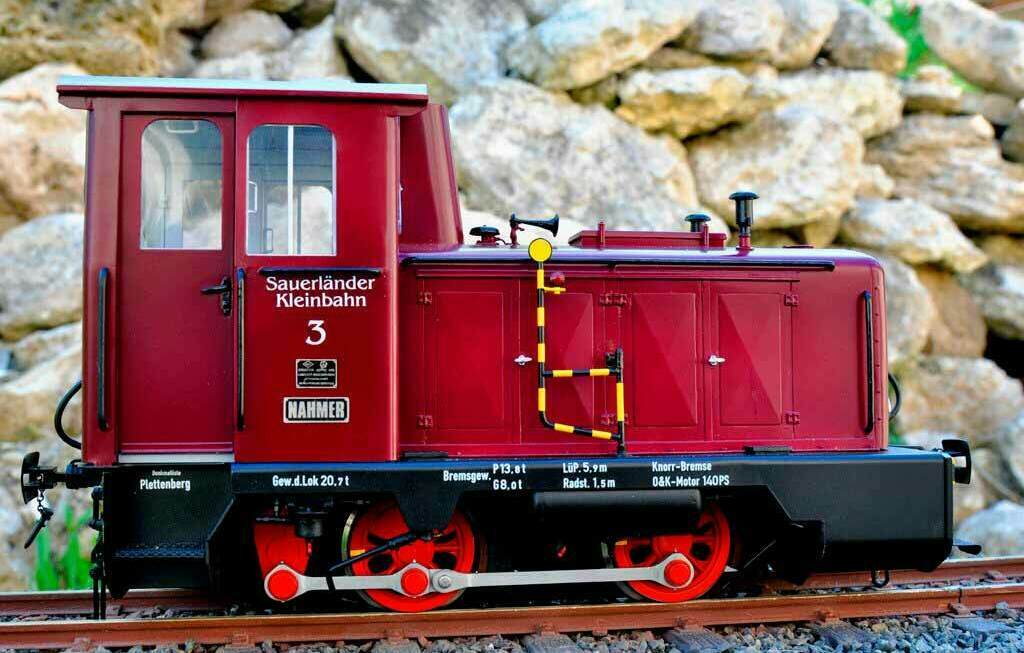Модель узкоколейного локомотива Sauerländer Kleinbahn 3 на рельсах Красная масштабная модель дизельного локомотива Sauerländer Kleinbahn 3 на узкоколейной железной дороге, вид сбоку крупным планом