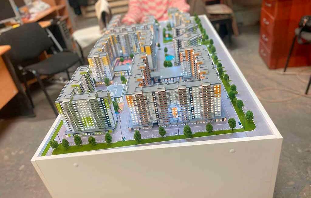 Макет жилого комплекса и зданий, выполненный на заказ для выставки Детализированный архитектурный макет многоэтажного жилого комплекса: модели домов, дворы, парковка и озеленение на подставке в офисе, выполненный на заказ для выставки