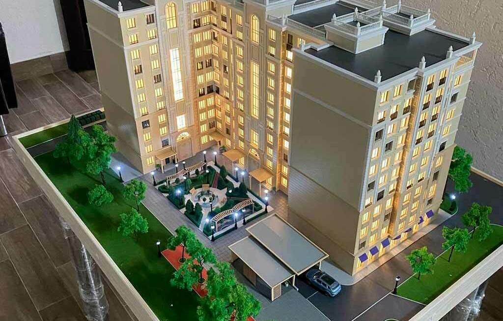 Архитектурный макет элитного жилого многоэтажного здания, выполненный на заказ для выставки Детализированный архитектурный макет многоэтажного жилого комплекса с освещённым двором, парковкой, дорожками и зелёными насаждениями на столе в офисе