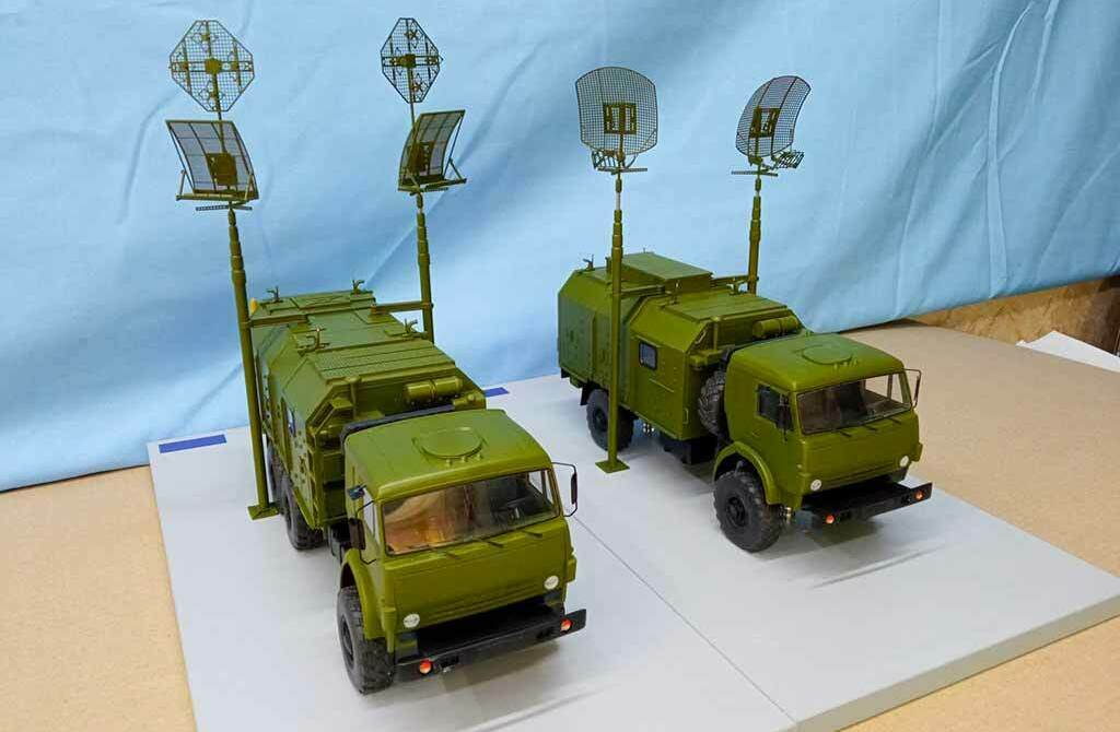 Масштабная модель военной радиолокационной станции на шасси грузовика Камаз Две зелёные масштабные модели военных радиолокационных машин на базе грузовиков с поднятыми антеннами, установленные на подставке на фоне голубой ткани